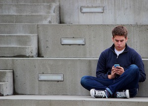 Juvenile Crimes Attorney in Los Angeles County Teenage Boy Sitting with His Legs Crossed on Steps Outside Looking at His Phone